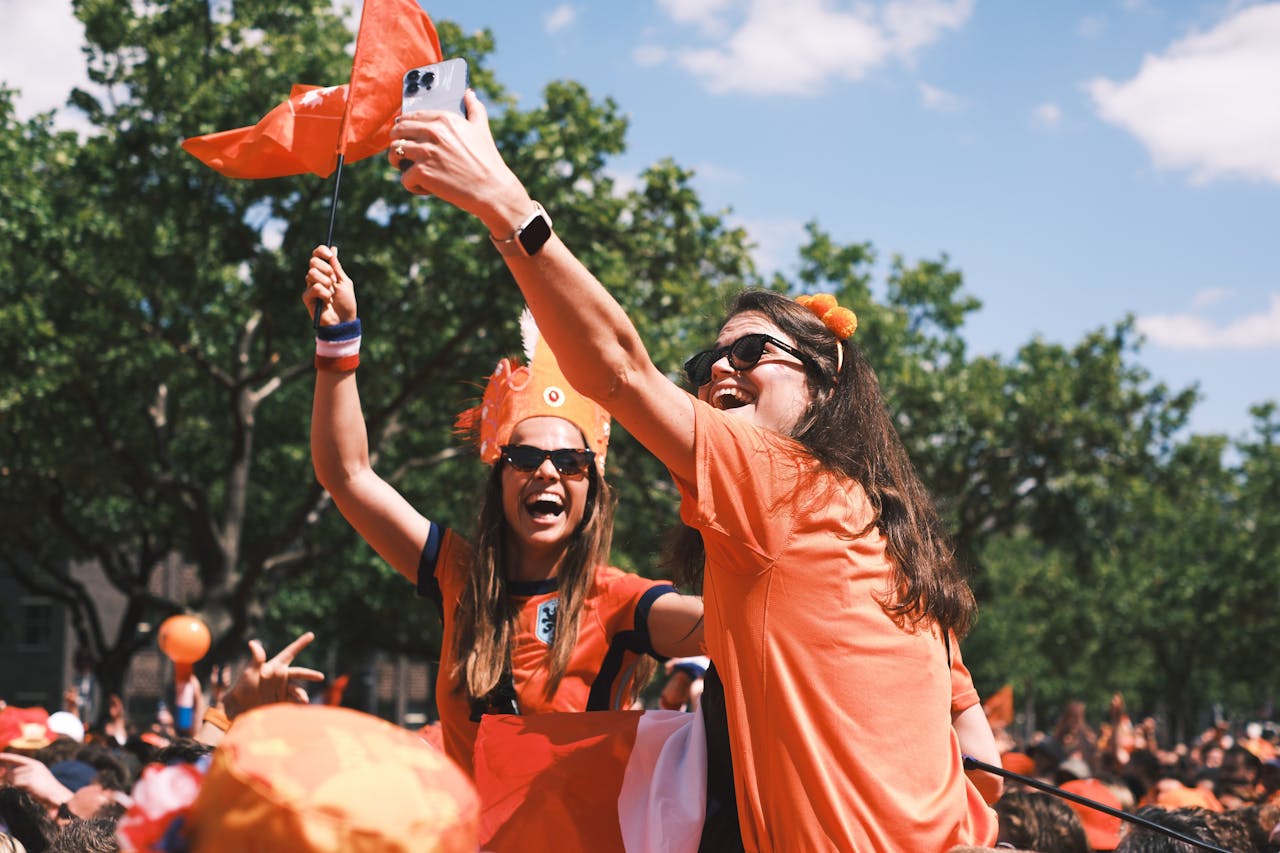 Excited Dutch football fans take selfies and celebrate outdoors wearing orange, waving flags.